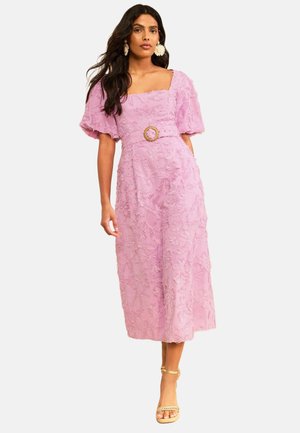 Žena oblečená v textúrovaných fialových midi šatách s nariasenými rukávmi a opaskom v páse, doplnená svetlo béžovými sandálmi s remienkom okolo členku a veľkými kvetinovými náušnicami.