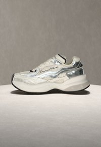 Άσπρο και ασημένιο chunky sneaker με κορδόνια μπροστά και μαύρες λεπτομέρειες στη σόλα, εμφανίζεται σε ανοιχτή επιφάνεια με απλό φόντο.