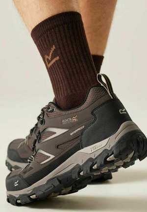 Zapato de senderismo impermeable marrón con suela resistente y detalles en negro, usado con calceta acanalada marrón oscuro en un pie elevado.