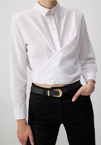 Camicia bianca con design avvolgente, dotata di colletto e maniche lunghe. Indossata con pantaloni neri e una cintura nera, arricchita da una fibbia d'oro.