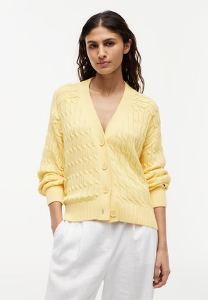 Femme portant un cardigan boutonné en maille torsadée jaune pâle avec les manches retroussées et un pantalon blanc, debout devant un fond clair uni.