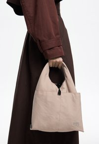 Torba w kolorze beżowym o miękkiej fakturze, z zaokrągloną górną rączką, czarnym zamkiem błyskawicznym i wyszywanym logo, trzymana w dłoni obok brązowego ubrania.