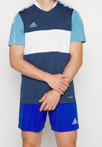 Adidas sportströja har horisontella ränder i marinblått, ljusblått och vitt. Tillverkad av andningsbart material med V-ringning och korta ärmar.