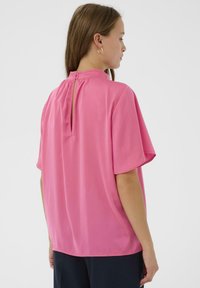 Top rosa con mangas cortas, textura suave, diseño en la espalda tipo ojal y cierre de botón. Corte simple y relajado con un dobladillo redondeado.