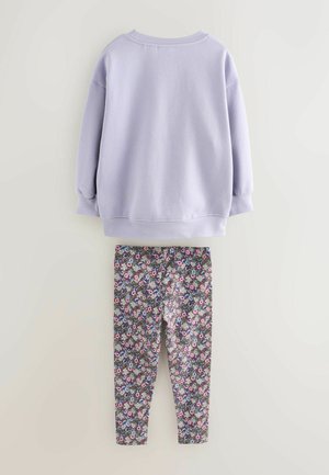 Hanorac de culoare lavandă deschisă cu mâneci lungi, purtat deasupra unei colanți negri ce prezintă un model floral colorat, cu nuanțe florale multiple pe un fundal închis.