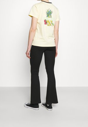 T-shirt jaune clair avec un graphique coloré de fruits dans le dos, associé à un pantalon évasé noir et des baskets noires.