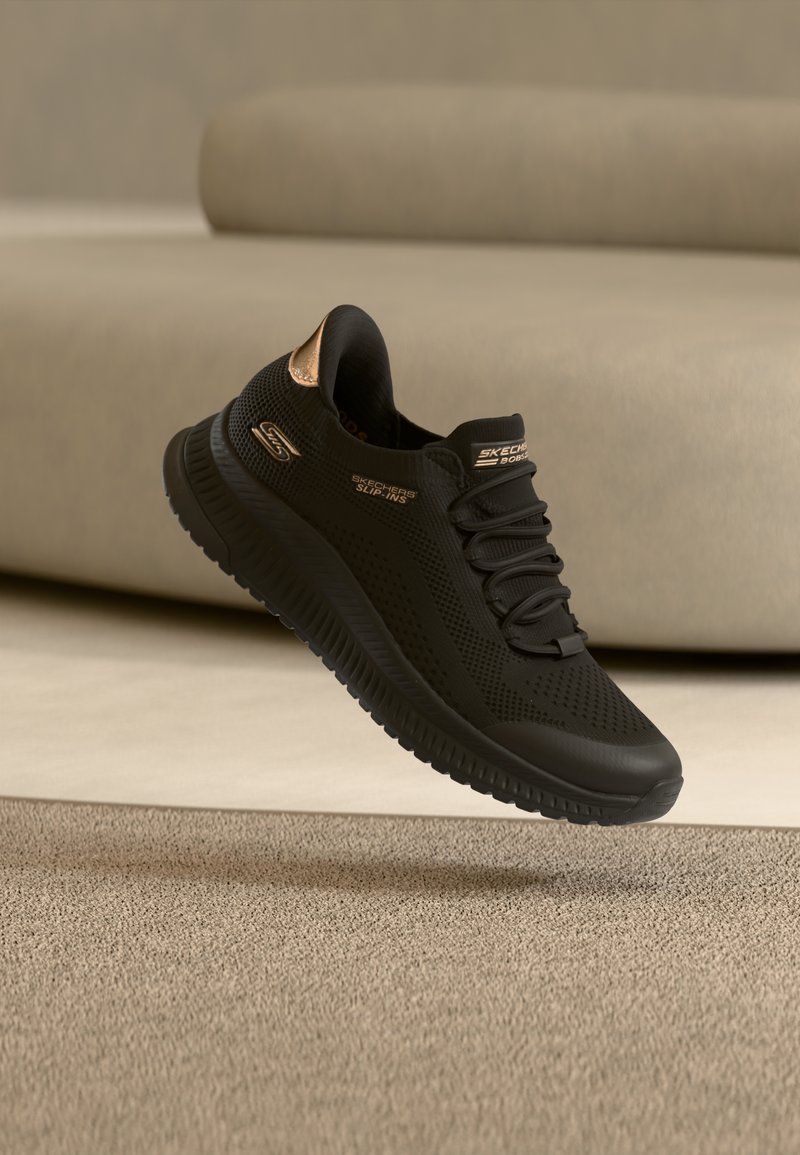 Baskets Skechers noires à enfiler avec dessus en mesh et accents en or rose, flottant au-dessus d'un tapis beige devant un canapé beige.