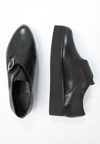 Une paire de chaussures à talons plateforme en cuir noir avec un détail de boucle en argent, l'une montrant la vue de dessus, l'autre la vue de profil sur fond blanc.