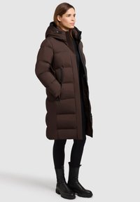 Giacca imbottita marrone con cappuccio, caratterizzata da un design trapuntato, tasche laterali e una silhouette lunga. Indossata con leggings neri e stivali.