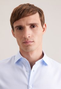 Jasnoniebieska koszula na guziki wykonana z fakturowanej tkaniny z klasycznym kołnierzykiem. Posiada pojedynczą kieszeń na piersi oraz subtelne przeszycia.