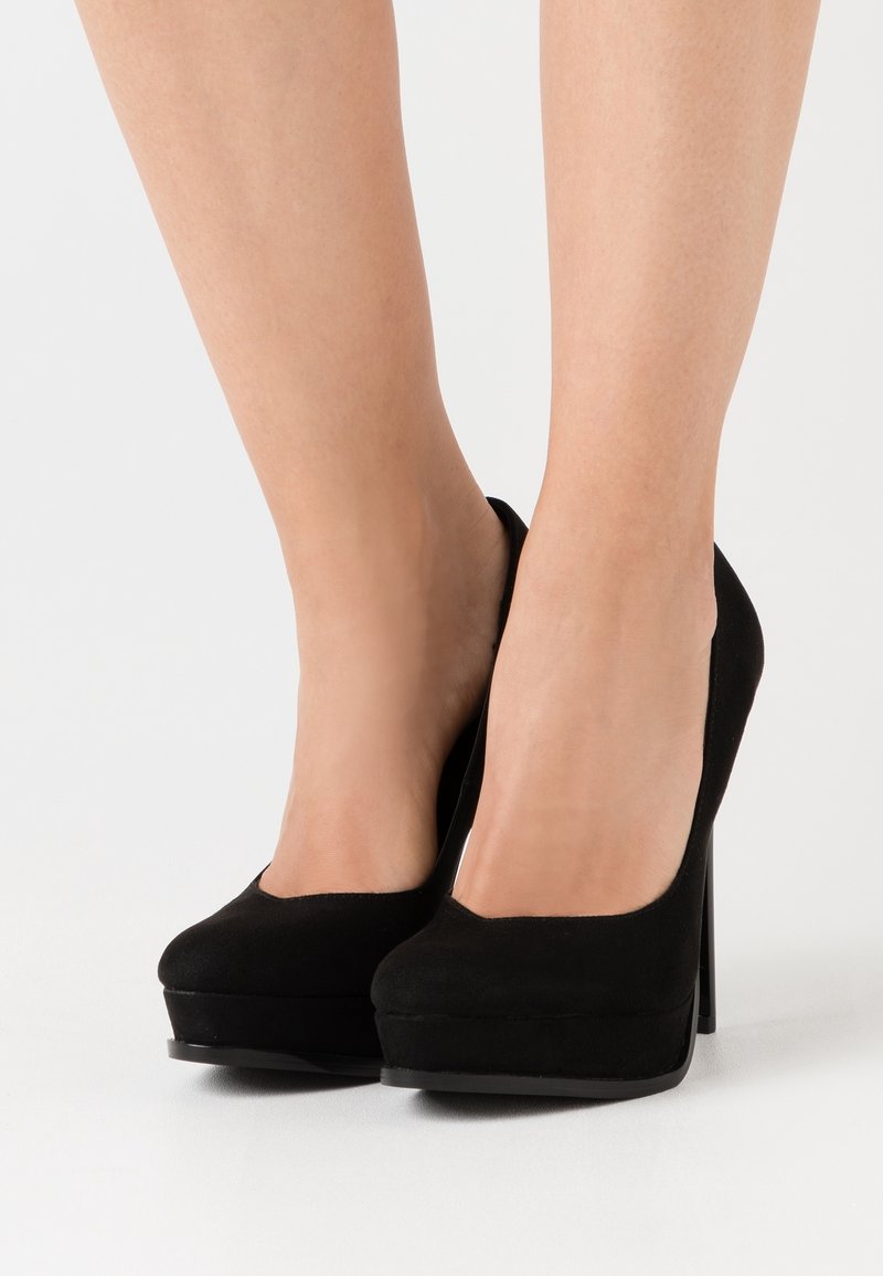 Jambes portant des escarpins à plateforme en daim noir avec bouts arrondis sur fond blanc.