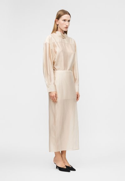 Femeie purtând o rochie lungă din satin bej pal, cu mâneci lungi și guler înalt, asortată cu pantofi negri cu vârf ascuțit și toc mic, stând pe lângă.