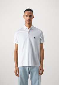 Polo shirt bianco a maniche corte con colletto a tre bottoni, dotato di un piccolo logo blu navy sul petto; materiale di cotone liscio e vestibilità rilassata.