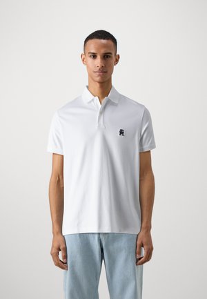 Polo shirt bianco a maniche corte con colletto a tre bottoni, dotato di un piccolo logo blu navy sul petto; materiale di cotone liscio e vestibilità rilassata.