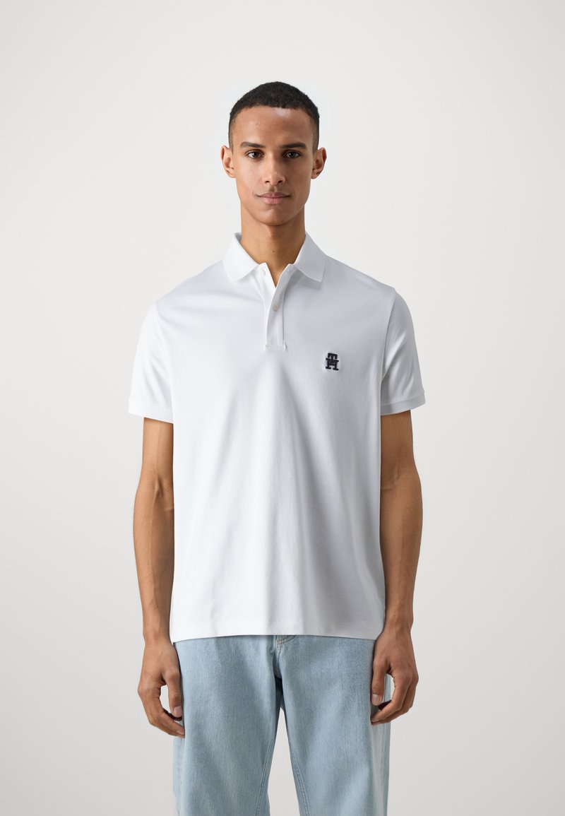 Polo shirt bianco a maniche corte con colletto a tre bottoni, dotato di un piccolo logo blu navy sul petto; materiale di cotone liscio e vestibilità rilassata.