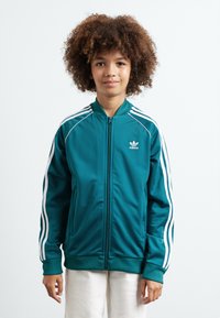 Tirkizna jakna na zip s bijelim trostrukim naglascima na rukavima, rebrastim manžetama i rubom, te malim bijelim Adidas logotipom na prsima.