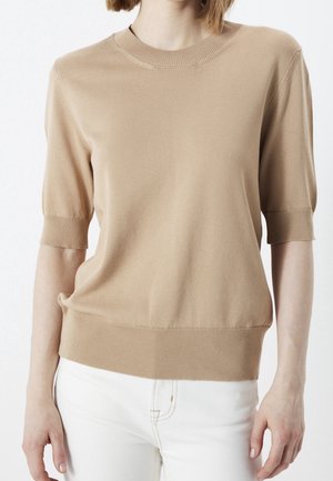 Beige kortærmet striktop med ribbet manchet og kant båret med hvide bukser mod en ensfarvet baggrund.