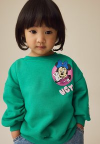 Grøn sweatshirt med et rundt Minnie Mouse grafik og "HEY" tekst; har ribbede mansjetter og en afslappet pasform. Denim shorts synlige forneden.