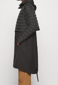 Chaqueta negra acolchada con diseño corto, sección inferior larga en negro, cierre de cremallera y bolsillos laterales. Cuenta con capucha y textura acolchonada.