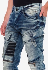 Blauwe denim cargobroek met een vervaagde afwerking, meerdere zakken, ritsen en gestructureerde accenten. Voorzien van een riemlus en een knoopsluiting.
