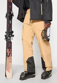 Bež hlače za smučanje z črnimi detajli, ujemajoče se s črnimi smučarskimi čevlji in smučjo. V roki ima tudi rokavice in smučarske očala.