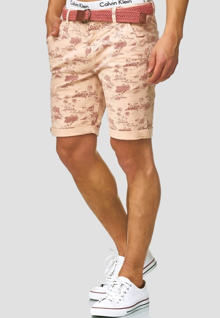 Homme portant un short beige avec un imprimé nature rouge, des revers roulés, une ceinture tissée rouge, une ceinture taille blanche Calvin Klein et des baskets blanches.