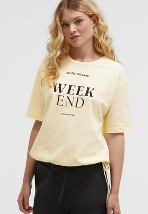 Blonde Frau trägt ein hellgelbes T-Shirt mit dem Aufdruck "SENSES. THE LABEL WEEK END NOTHING BUT YOU." und schwarze Hosen mit Tunnelzug.