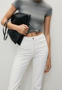 Top corto gris con textura acanalada anudado en el frente, combinado con jeans blancos ajustados. Se sostiene un bolso negro tipo plegable bajo el brazo.