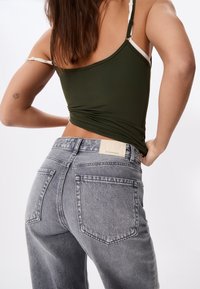 Femme portant un caraco vert foncé avec des bretelles blanches et un jean gris taille haute, montrant le dos et un tatouage sur le bras gauche.