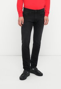 Črne denim hlače z ožjim krojem, petimi žepi in gumbom za zapiranje, v kombinaciji s črnimi natikači in svetlo rdečo majico z dolgimi rokavi.