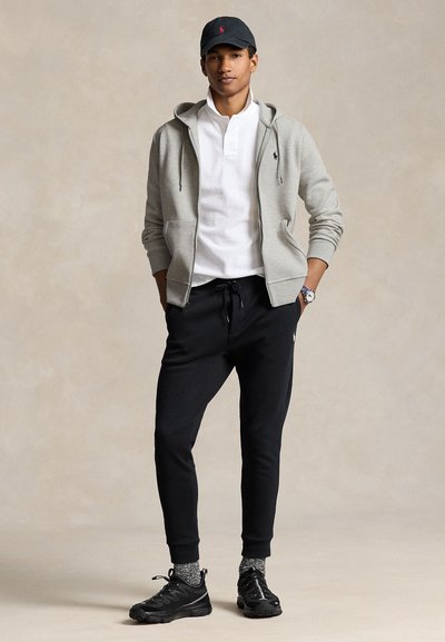 Ung mand iført en grå hættetrøje, hvid polo shirt, sorte joggingbukser, sorte sneakers, en sort kasket og et armbåndsur står med hænderne i lommerne.