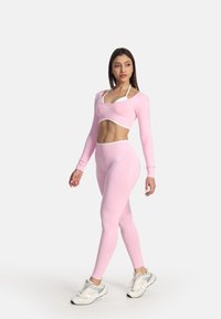Femme portant un crop top à manches longues rose et des leggings avec un soutien-gorge de sport blanc et des baskets blanches, debout sur un fond uni.