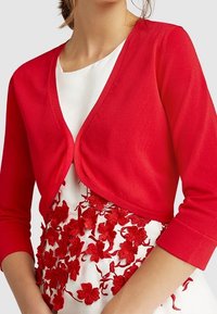 Rød strik cardigan med trekvartærmer og V-udskæring, båret over en hvid kjole med røde blomsterbroderier.