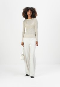 Lys beige ribbet langermet topp kombinert med hvite vide bukser. Modellen holder en liten hvit veske. Svarte spisse sko.