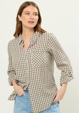 Frau trägt ein beige-braunes kariertes Hemd mit aufgekrempelten Ärmeln und hellblauen Jeans, blickt zur Seite.