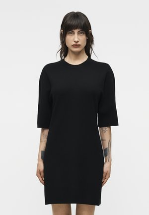 Γυναίκα με σκούρα μαλλιά και τατουάζ, που φοράει απλό μαύρο φόρεμα μήκους μέχρι το γόνατο με μανίκια μέχρι τους αγκώνες, στέκεται μπροστά σε ανοιχτόχρωμο φόντο.