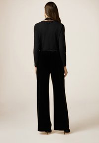 Zwarte fluwelen jumpsuit met lange mouwen, een wijd uitlopende ontwerp en een gestructureerd oppervlak. Afgewerkt met een strikdetail op de achterkant van de halslijn.