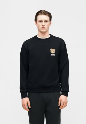 Ung mand iført en sort sweatshirt med en lille bamse-logo og teksten "Moschino Underbear" på brystet.