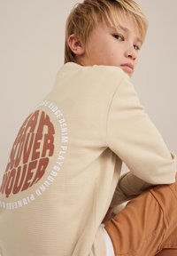 Chemise texturée beige clair avec imprimé graphique rond au dos. Accentuée par des lettres marron foncé et des détails blancs. Coupe décontractée.