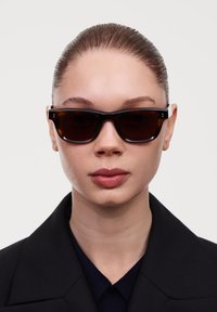Ochelari de soare tortoise dark, cu o ramă rectangulară groasă, lenti întunecate și accente argintii pe timpane. Purtați cu un palton negru.