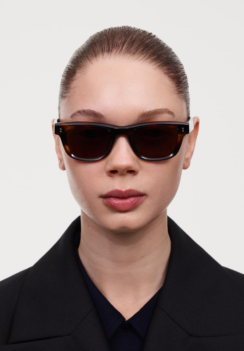 Ochelari de soare tortoise dark, cu o ramă rectangulară groasă, lenti întunecate și accente argintii pe timpane. Purtați cu un palton negru.