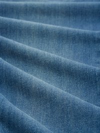 Tissu en denim avec une texture lisse ; couleur bleu moyen avec des variations subtiles. Présente un motif de tissage diagonal et un léger éclat dû à la finition du tissu.