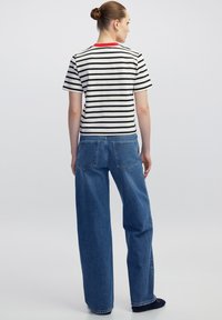 Mujer de piel clara y cabello recogido en un moño, de espaldas, vistiendo una camiseta a rayas black y white con collar rojo y pantalones vaqueros azul holgados.