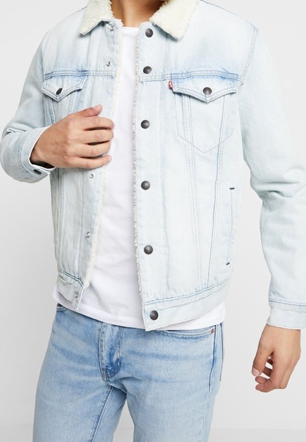 Veste en denim bleu clair avec col en polaire blanche portée sur un t-shirt blanc, associée à un jean bleu clair, personne ajustant le devant de la veste.
