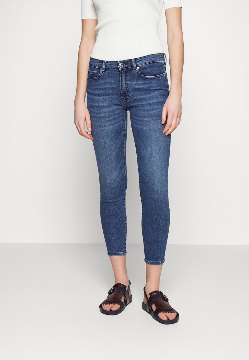 Personne portant un jean bleu cropé ajusté avec un haut blanc à manches courtes et des sandales ouvertes marron foncé avec des détails de boucle.