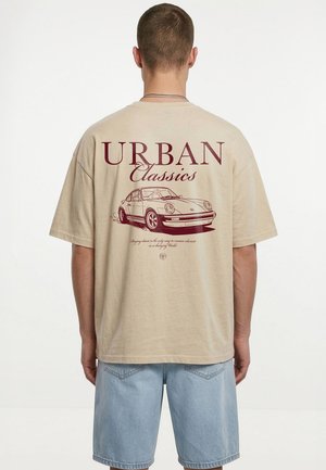 Mann trägt ein beige T-Shirt mit einer Skizze eines Oldtimers und dem Schriftzug „Urban Classics“ auf dem Rücken, kombiniert mit hellblauen Denim-Shorts.