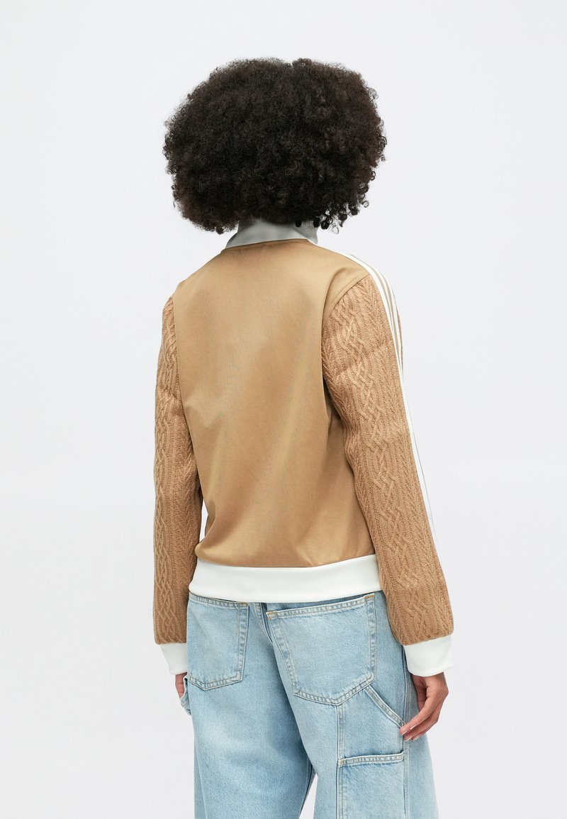 Braune Strickjacke mit Reißverschluss und strukturierten Ärmeln sowie weißen Akzenten. Verfügt über einen glatten Stoffkörper und gerippte Bündchen. Hellblaue weit geschnittene Jeans sind sichtbar.