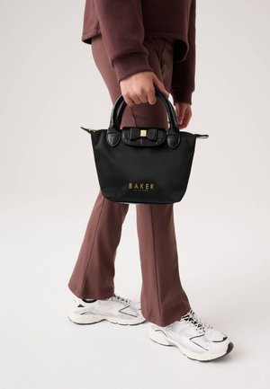 Una persona con pantalones de color malva y zapatillas blancas sostiene un bolso negro de Ted Baker con letras doradas sobre un fondo liso.