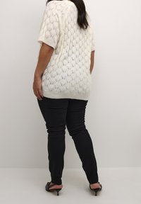 Biały sweter dzianinowy z teksturowanym wzorem w diamenty, krótki rękaw i zaokrąglony dół, zestawiony z czarnymi spodniami slim-fit i sandałami na obcasie z odkrytymi palcami.