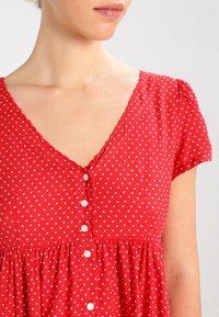 Röd klänning med korta ärmar, v-ringning, vitt stjärnmönster och knappstängning fram. Mjuk tyg med rynkad nedre kant.